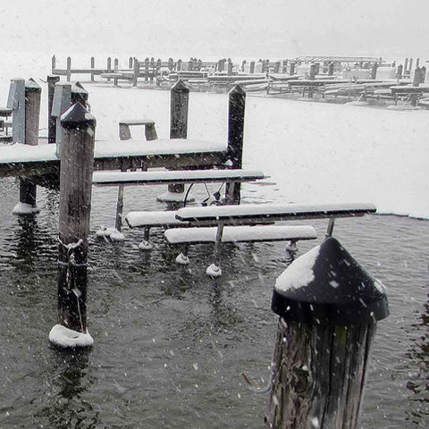 Snow-covered dock with benches and posts in a body of water during a snowfall.